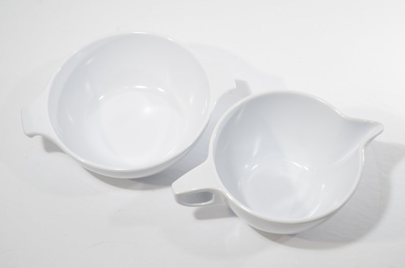 May include: Two white ceramic bowls, one with a handle and the other with a pouring spout. Both bowls are empty and appear to be made of a smooth, glossy material.