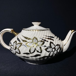 Könnte beinhalten: Eine weiße Teekanne mit goldfarbenem und schwarzem Blumendesign. Die Teekanne hat einen runden Korpus und einen Ausguss auf der rechten Seite. Der Henkel befindet sich auf der linken Seite.