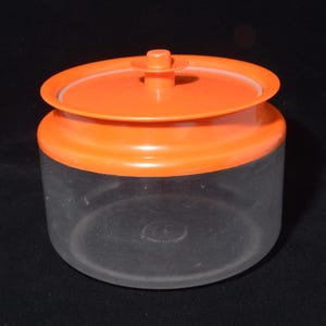 May include: A clear plastic container with an orange lid. The lid has a circular design with a small handle on top. The container is cylindrical and sits on a dark surface.