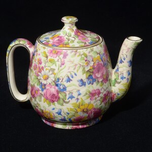 Puede incluir: Una tetera de cerámica con una base de color crema adornada con un vibrante patrón floral multicolor que incluye rosas rosadas, margaritas amarillas y flores azules en forma de campana. La tetera incluye una tapa a juego, un asa curva y un pico.
