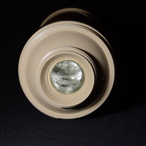 May include: A beige-colored, cylindrical thermos bottle with a wide, circular top and a narrower base. The interior is visible, reflecting light. The thermos is set against a dark background, highlighting its shape and color.