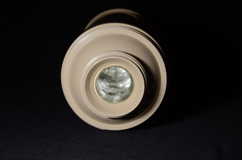 May include: A beige-colored, cylindrical thermos bottle with a wide, circular top and a narrower base. The interior is visible, reflecting light. The thermos is set against a dark background, highlighting its shape and color.