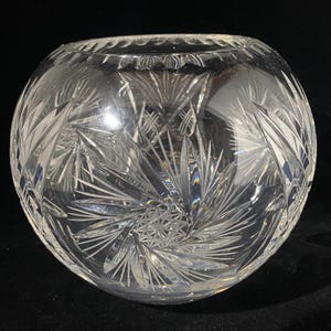 May include: A clear, round crystal bowl with intricate starburst and leaf-like patterns etched into the glass. The bowl has a wide opening and a decorative rim, reflecting light and creating a sparkling effect. The bowl is likely used for decorative purposes.