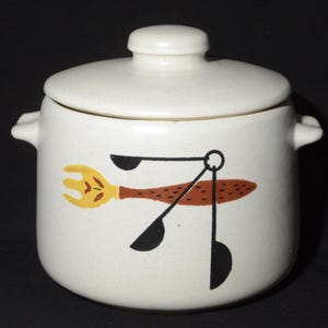 May include: Cream ceramic jar with lid, decorated with a fork, spoon, and measuring spoons in yellow, brown, and black. The jar has two small handles and a matching lid with a small knob. A kitchenware item.
