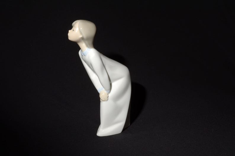 May include: A porcelain figurine of a girl in a white dress with blue trim, looking up and to the left.