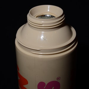 May include: A beige insulated bottle with a screw-on top. The top has a wide opening and a silver interior. The bottle's body is cylindrical, and the design includes orange and pink lettering. The background is black.