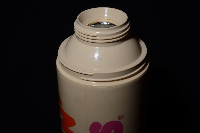 May include: A beige insulated bottle with a screw-on top. The top has a wide opening and a silver interior. The bottle's body is cylindrical, and the design includes orange and pink lettering. The background is black.