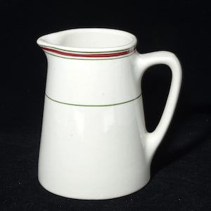 May include: White ceramic pitcher with a handle. The pitcher has a red and green stripe around the rim and a green stripe around the body. The pitcher is approximately 6 inches tall.