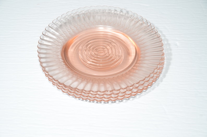 Puede incluir: Un conjunto de platos de vidrio rosa apilados. Cada plato presenta un borde acanalado y un centro circular y estriado. Los platos son de un tono rosa suave y est&aacute;n apilados. Se presentan sobre una superficie blanca.