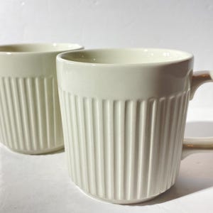 May include: Two white ceramic mugs with vertical ridges. The mugs have a simple design and are perfect for everyday use.