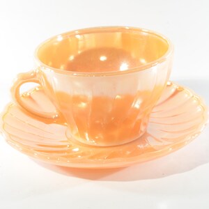 May include: A vintage peach-coloured teacup and saucer set. The cup and saucer are made of iridescent glass and have a delicate, scalloped edge.