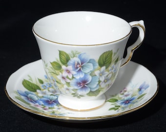 Könnte beinhalten: Weißes Tee-Set mit einer Blumengestaltung mit blauen, lila und weißen Blumen und grünen Blättern. Die Tasse und die Untertasse haben einen goldenen Rand. Ein klassisches Teeservice.
