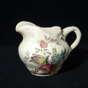 Peut inclure: Crémier en céramique crème avec un motif floral. Le crémier présente un bec verseur incurvé et une anse. Le motif floral comprend des fleurs roses, bleues et rouges avec des feuilles vertes. Le crémier est probablement vintage ou antique.