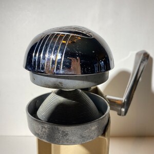 May include: A vintage chrome and silver manual citrus juicer with a lever handle. The juicer has a ribbed, conical reamer and a chrome top with a raised design.