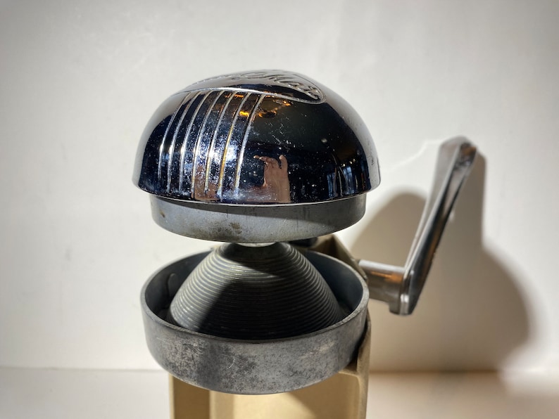 May include: A vintage chrome and silver manual citrus juicer with a lever handle. The juicer has a ribbed, conical reamer and a chrome top with a raised design.