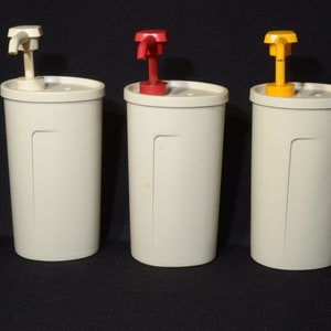 Peut inclure: Trois distributeurs cylindriques crème avec des pompes de couleurs différentes. Le premier a une pompe blanche, le second une pompe rouge et le troisième une pompe jaune. Les distributeurs sont sur fond noir.