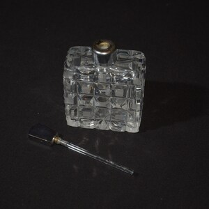 May include: A clear glass bottle with a square shape and a silver top. The bottle has a textured surface and is sitting on a black surface. A silver metal dropper is next to the bottle.