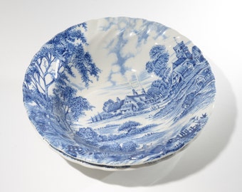 May include: A blue and white ceramic bowl with a scalloped rim. The bowl features a transferware design of a countryside scene with houses, trees, and a church.