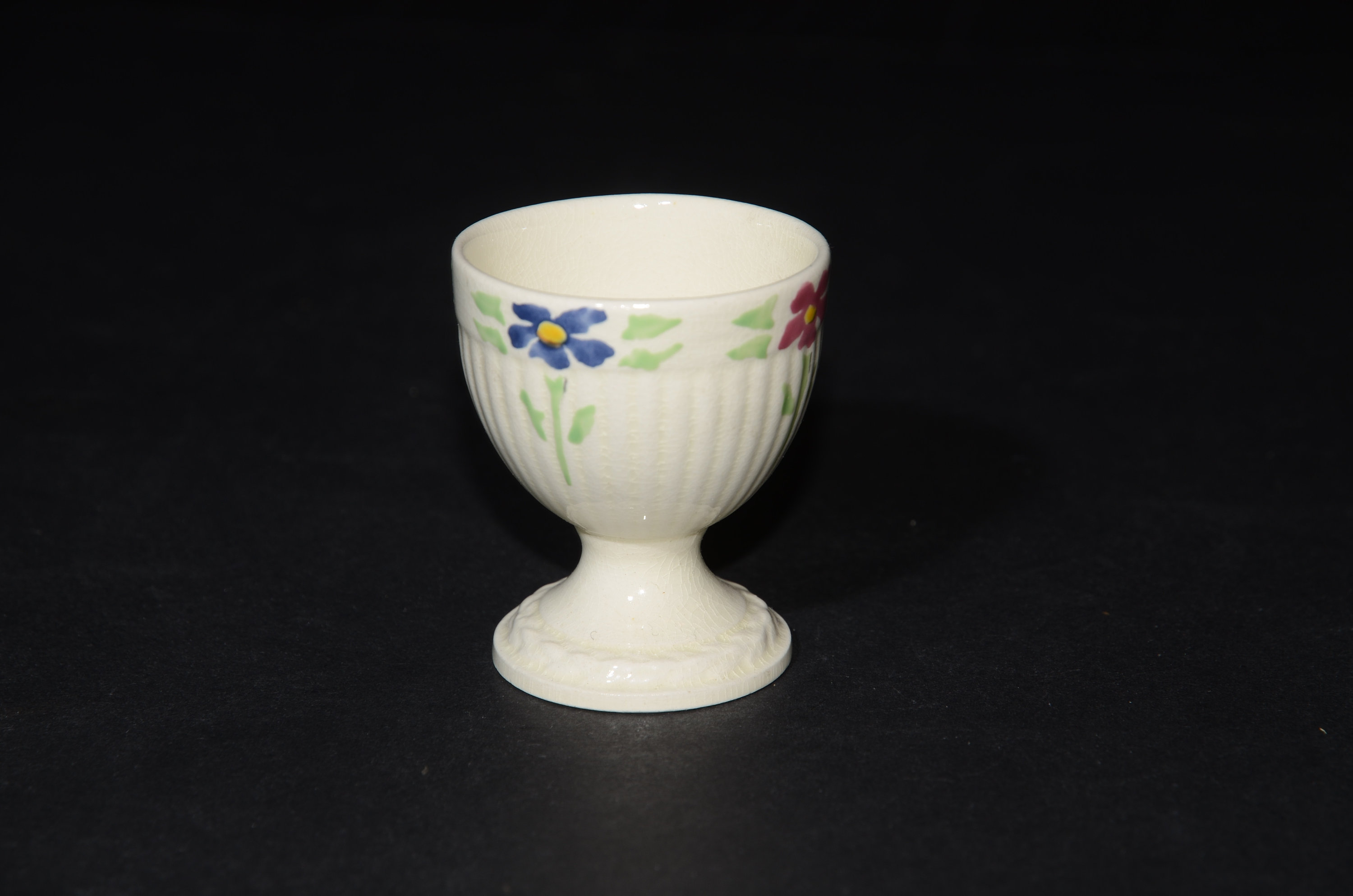 May include: White ceramic egg cup with a floral design. The cup has a fluted base and a rim with blue, red, and green flowers.