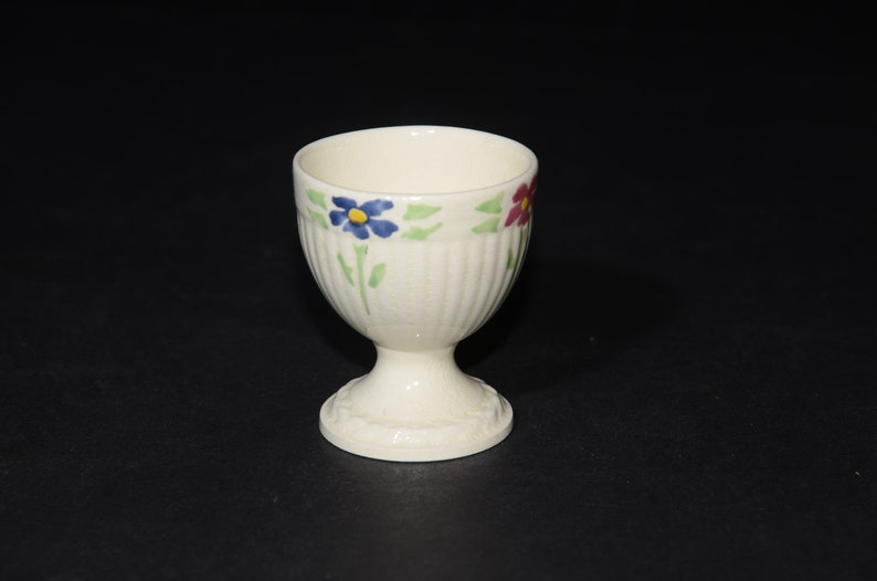 May include: White ceramic egg cup with a floral design. The cup has a fluted base and a rim with blue, red, and green flowers.