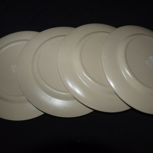 May include: Four beige ceramic dinner plates stacked on top of each other. The plates have a slightly raised rim and a smooth, glossy finish.