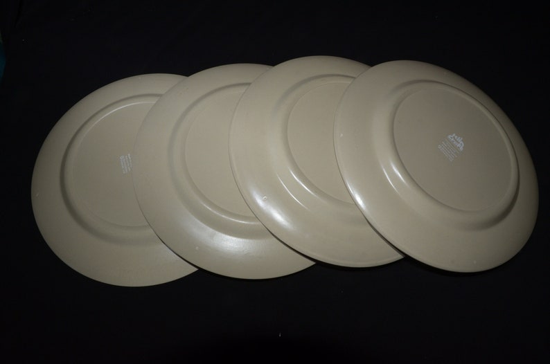 May include: Four beige ceramic dinner plates stacked on top of each other. The plates have a slightly raised rim and a smooth, glossy finish.