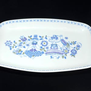 May include: A rectangular white ceramic serving platter featuring a blue and purple illustration of two figures surrounded by stylized flowers and a bird. The rim is decorated with a repeating geometric pattern in blue, creating a border.