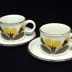 May include: Two ceramic coffee cups and saucers. The cups and saucers are white with a brown rim. Each cup features a yellow floral design with brown leaves. The cups have a handle.