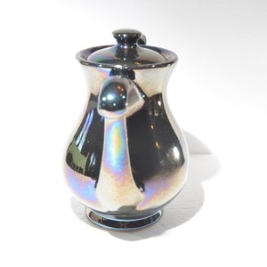 May include: A silver iridescent ceramic creamer with a lid and a handle. The creamer has a pearlescent finish.