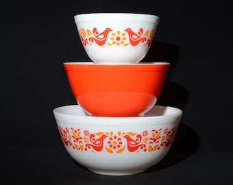 May include: A stack of three mixing bowls. The top and bottom bowls are white with a repeating orange and yellow pattern of stylized birds and flowers. The middle bowl is solid bright orange. All three bowls have a smooth, rounded shape.