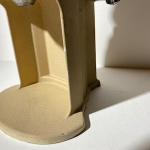 May include: Close-up of the base of a beige electric coffee grinder. The base is made of a textured material and has a rounded shape.