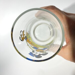 May include: Clear glass with a gold rim featuring colorful cartoon characters Mickey Mouse and Goofy. The glass is round and appears to be empty, with the characters printed on the side. The image is taken from a top-down perspective.