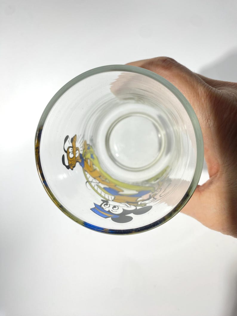 May include: Clear glass with a gold rim featuring colorful cartoon characters Mickey Mouse and Goofy. The glass is round and appears to be empty, with the characters printed on the side. The image is taken from a top-down perspective.