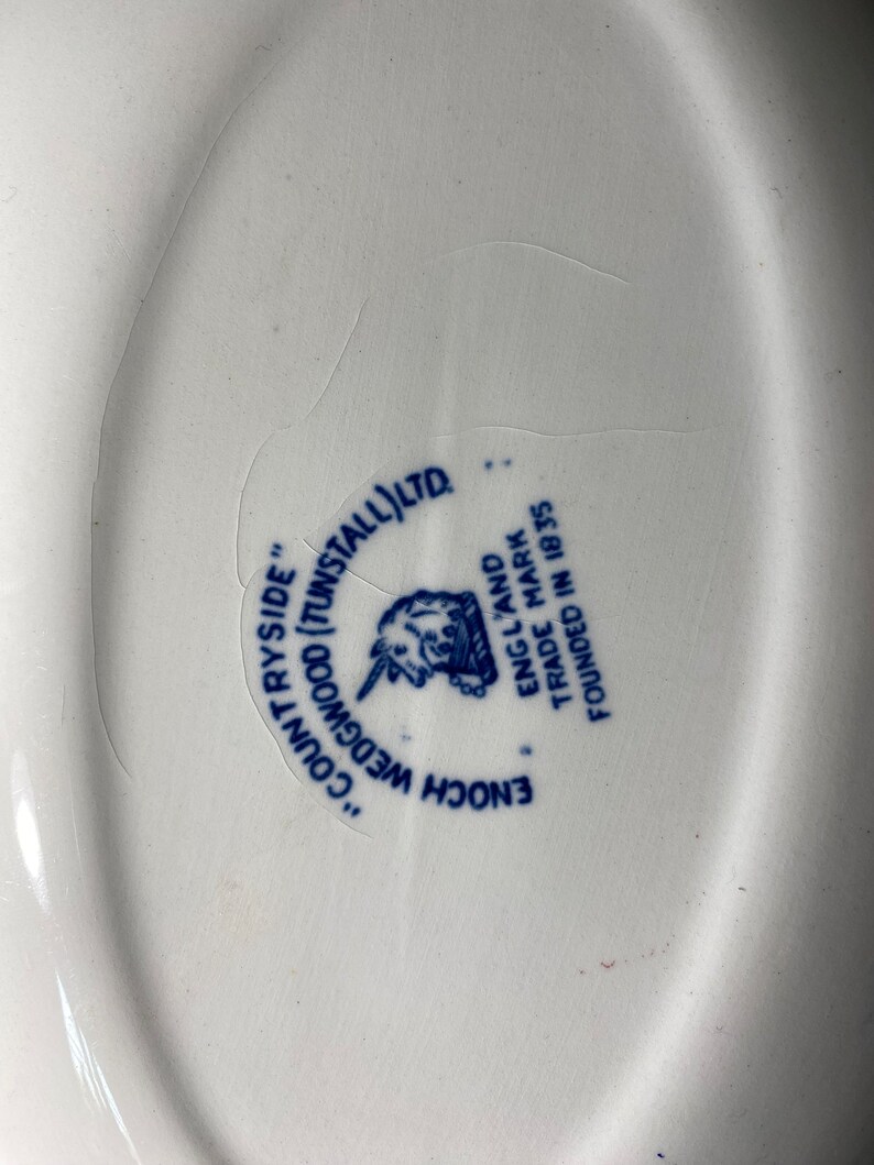 Op de afbeelding: Een close-up van een wit keramiek bord met een blauw en wit logo. Het logo luidt "Enoch Wedgwood (Tunstall) Ltd." en "Engeland Handelsmerk Gevestigd in 1835".