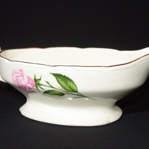 Peut inclure: Saucière en porcelaine blanche avec un motif de rose rose et de feuilles vertes. La saucière a un bord doré et une anse.
