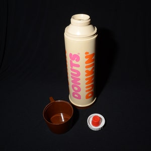 May include: A vintage Dunkin' Donuts thermos with a brown cup and a red and white lid. The thermos is beige with the words "DONUTS" in pink and "DUNKIN'" in orange. The cup is brown.