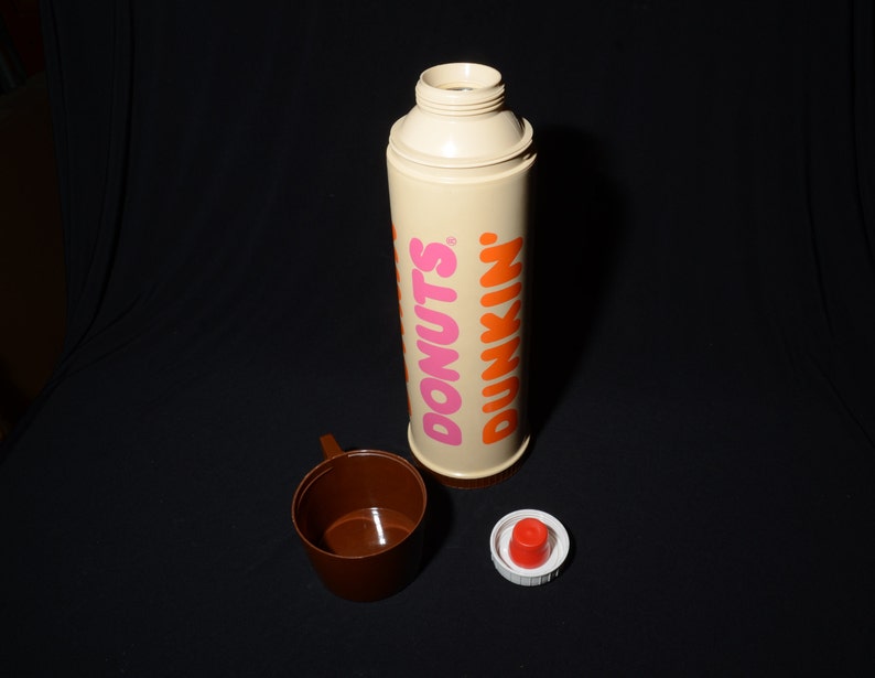 May include: A vintage Dunkin' Donuts thermos with a brown cup and a red and white lid. The thermos is beige with the words "DONUTS" in pink and "DUNKIN'" in orange. The cup is brown.