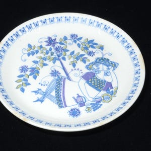Puede incluir: Plato de cerámica blanco con una ilustración azul y morada de una persona relajándose bajo un árbol. El plato tiene un borde floral azul y un diseño a juego en el borde. También se representa una tetera y una taza.