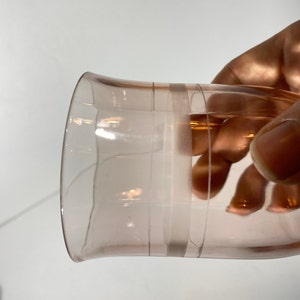 May include: A clear pink glass with a wide rim and a slightly flared base. The glass has two horizontal lines around the middle.