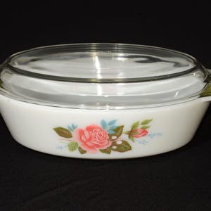 May include: White oval casserole dish with a clear glass lid. The dish features a floral design with pink roses, green leaves, and blue accents. The handles are white with a green trim.