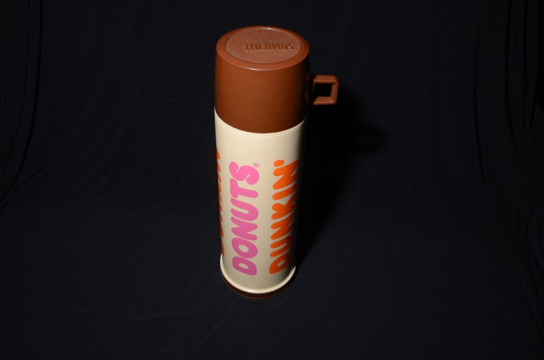 May include: A vintage brown and off-white Thermos brand travel mug. The mug features the Dunkin' Donuts logo in pink and orange. The lid and handle are brown. The mug is cylindrical and stands upright against a black background.