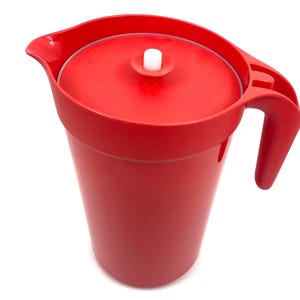 Puede incluir: Una jarra de plástico de color rojo brillante que incluye una tapa a juego con un pequeño pomo cilíndrico blanco en el centro. La jarra cuenta con un pico vertedor ancho en un lado y un asa integrada resistente en el lado opuesto.