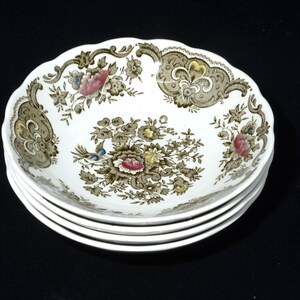 Può includere: Una pila di quattro ciotole in ceramica bianca con bordo smerlato. Ogni ciotola presenta un motivo floreale nei toni del marrone, rosa e blu. Il design include fiori e fogliame, creando un'estetica vintage. Lo sfondo è nero.