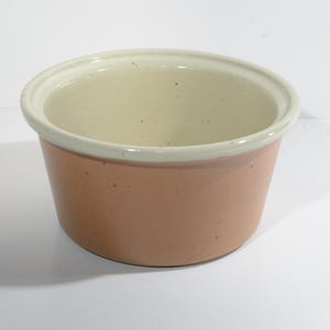 May include: A round, ceramic bowl with a light peach exterior and a cream-colored interior. The rim is also cream-colored. The bowl is likely used for serving or food preparation. The bowl is sitting on a white surface.