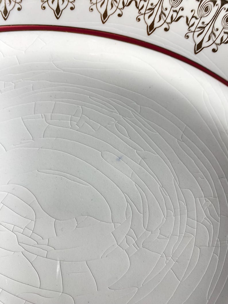 May include: Close-up of a white ceramic plate with a cracked, crazed glaze. The plate has a red border and a decorative brown pattern around the rim. The surface shows fine cracks.