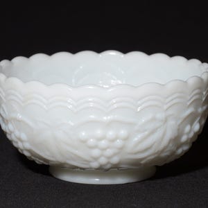 K&ouml;nnte beinhalten: Eine wei&szlig;e Milchglasschale mit gewelltem Rand und Sockel. Die Schale hat ein gepr&auml;gtes Design aus Trauben und Bl&auml;ttern an der Au&szlig;enseite. Die Schale steht vor einem dunklen Hintergrund.