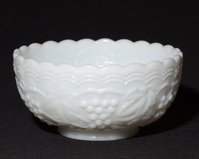 K&ouml;nnte beinhalten: Eine wei&szlig;e Milchglasschale mit gewelltem Rand und Sockel. Die Schale hat ein gepr&auml;gtes Design aus Trauben und Bl&auml;ttern an der Au&szlig;enseite. Die Schale steht vor einem dunklen Hintergrund.