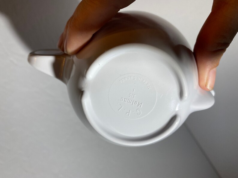 May include: White ceramic teapot with a handle and spout. The bottom of the teapot has the text "C.P. Melmac 1 qt. Made in Japan" printed on it.