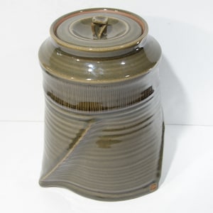 Könnte beinhalten: Eine grüne Keramikdose mit Deckel. Die Dose hat ein strukturiertes, wellenförmiges Design und einen braunen Rand.