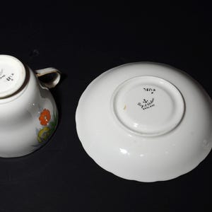 May include: White teacup and saucer set with floral design. The teacup has a handle and a floral pattern in orange, yellow, and green. The saucer is round with a scalloped edge. Both pieces have markings that say "England".